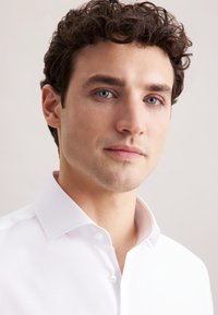 Witte button-up shirt met een puntige kraag, met een soepele textuur en een subtiele glans, gedragen door een man met kort, krullend haar en blauwe ogen.