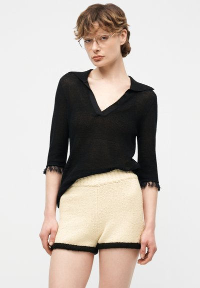 Femme portant des lunettes transparentes, un haut noir à manches trois-quarts avec col, et un short en maille crème avec bordure noire, debout devant un fond neutre.