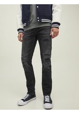 Hombre vistiendo jeans negros rasgados, zapatillas blancas y negras, sudadera con capucha gris y chaqueta universitaria azul marino con mangas blancas.