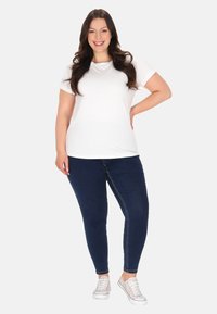 Hvid kortærmet t-shirt kombineret med mørkeblå skinny jeans. Modellen bærer afslappede hvide sneakers. Glat stof, løs pasform, enkelt design.