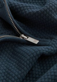 Tessuto testurizzato blu navy con motivo a nido d'ape, dotato di una cerniera argentata con tirante logo. Tessuto morbido che garantisce comfort e calore.