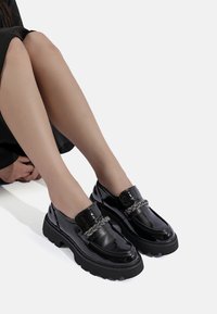 Mocassini in pelle patinata nera con suole chunky nere e accenti decorativi in catena metallica sulla parte superiore. Finitura liscia e lucida.