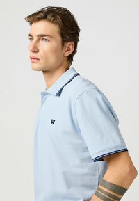 Ljusblå polo-tröja med en texturerad tyg. Har en marinrandig krage och en liten logotyp på vänster bröst. Kortärmad.