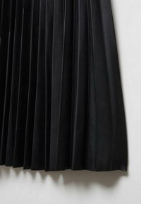 Jupe noire plissée en similicuir suspendue sur un fond blanc, montrant la texture et des plis verticaux nets.