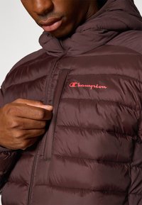 Burgundowa kurtka puchowa z kapturem, z pikowanym wzorem i czerwonym logo "Champion" na piersi. Kieszeń zapinana na zamek po lewej stronie.