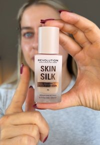 Revolution Skin Silk Luminous Serum Foundation i en klar glassflaske, med et rosa lokk og brun tekst. Størrelse: 23 ml, F5 nyanse.