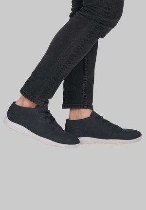 Personne portant des chaussures décontractées noires à lacets avec des semelles blanches et un jean foncé sur un fond gris uni.