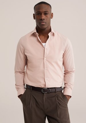 Mand iført lyserød skjorte med knapper over en hvid undertrøje, brune plisserede bukser og sort bælte, hænderne i lommerne, neutral baggrund.