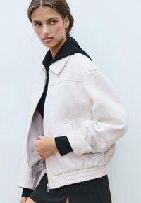 Veste bomber rose clair avec une texture pelucheuse, dotée d'une capuche noire, de poignets côtelés et d'une fermeture éclair à l'avant. Détails de poche simples.