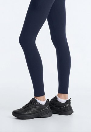 Legs wearing navy leggings and white ankle socks with black chunky athletic sneakers on a light background.