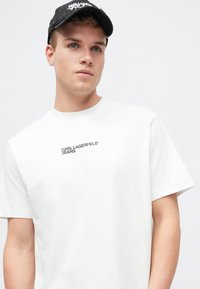T-shirt de algodão branca com a inscrição "KARL LAGERFELD JEANS" em cinza, com gola redonda e mangas curtas; modelo a usar uma chapéu preto.