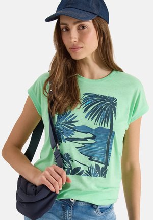 Femme portant un t-shirt vert avec un imprimé plage tropicale bleu, une casquette en denim bleue, un jean en denim et tenant un sac à bandoulière bleu marine.