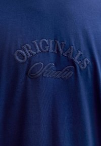 Camiseta de algodón azul marino con bordado texturizado que dice "ORIGINALS" sobre "Studio" en un tono contrastante. Textura de tejido suave.