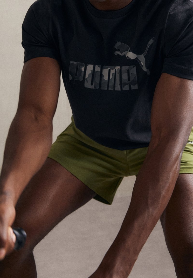 Černé sportovní tričko s maskáčovým logem Puma, spárované s olivově zelenými šortkami. Látka vypadá hladce, má přiléhavé rukávy a atletický střih.
