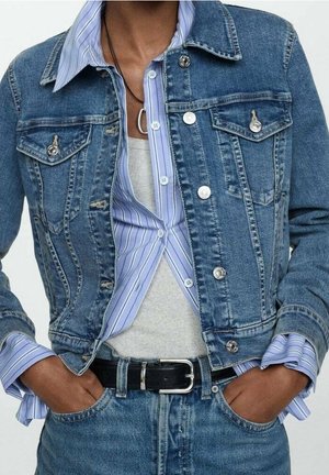 Person trägt eine blaue Jeansjacke über einem blau gestreiften Hemd mit Knöpfen und grauem Oberteil, mit Händen in den Taschen der blauen Jeans und einem schwarzen Gürtel.