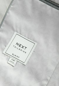 Forro de traje ajustado en gris claro con un patrón geométrico en relieve. Incluye una etiqueta blanca con "NEXT TAILORING" y detalles de tamaño.