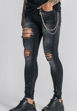 Person iført sorte skinny jeans med huller, en sølvkæde og sorte teksturerede sneakers, stående med den ene hånd i lommen.