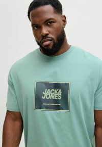 Miętowa bawełniana koszulka z nadrukiem z tekstem "JACK & JONES" umieszczonym w prostokątnym obramowaniu. Krótkie rękawy, okrągły dekolt.