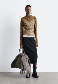Eine Frau trägt eine durchsichtige, senffarbene, blumige Bluse mit langen Ärmeln, einen schwarzen Midirock und schwarze Schnürstiefel. Sie hält eine olivgrüne Tote-Tasche mit einem grauen Fransenschal.