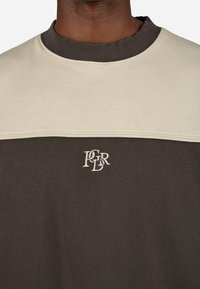 Svetr s barevným blokováním, který má béžovou horní část a hnědou dolní část. Obsahuje žebrovaný límec a vyšívaný detail loga.