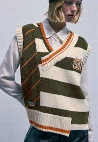 Wollmischung Strickweste in Grün, Creme und Orange gestreift, mit tiefem V-Ausschnitt und gesticktem Detail "1987". Strukturiert gerippter Saum.