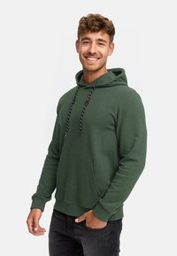 Grøn hoodie lavet af tekstureret stof, med en lomme foran, justerbare snore og en afslappet pasform. Sammen med mørke jeans.