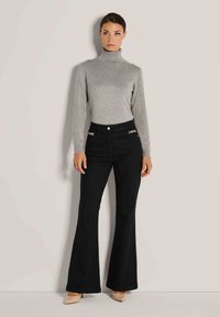 Vrouw in een grijze coltrui, zwarte hoge flarebroek met gouden kettingdetails, en beige puntige hakken staande tegen een beige muur.