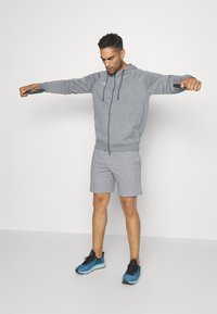 Grå atletisk hettegenser med kengurulomme, kombinert med matchende shorts. Modell iført blå og svarte joggesko, utfører en treningsposisjon.