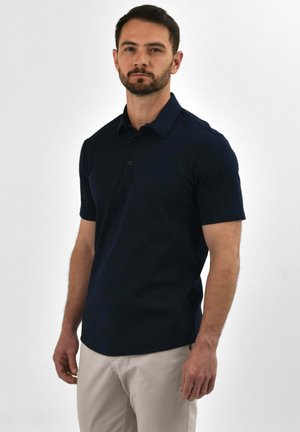 Man met kort donker haar en baard, gekleed in een marineblauw polohemd met korte mouwen en lichtbeige broek, staand tegen een effen witte achtergrond.