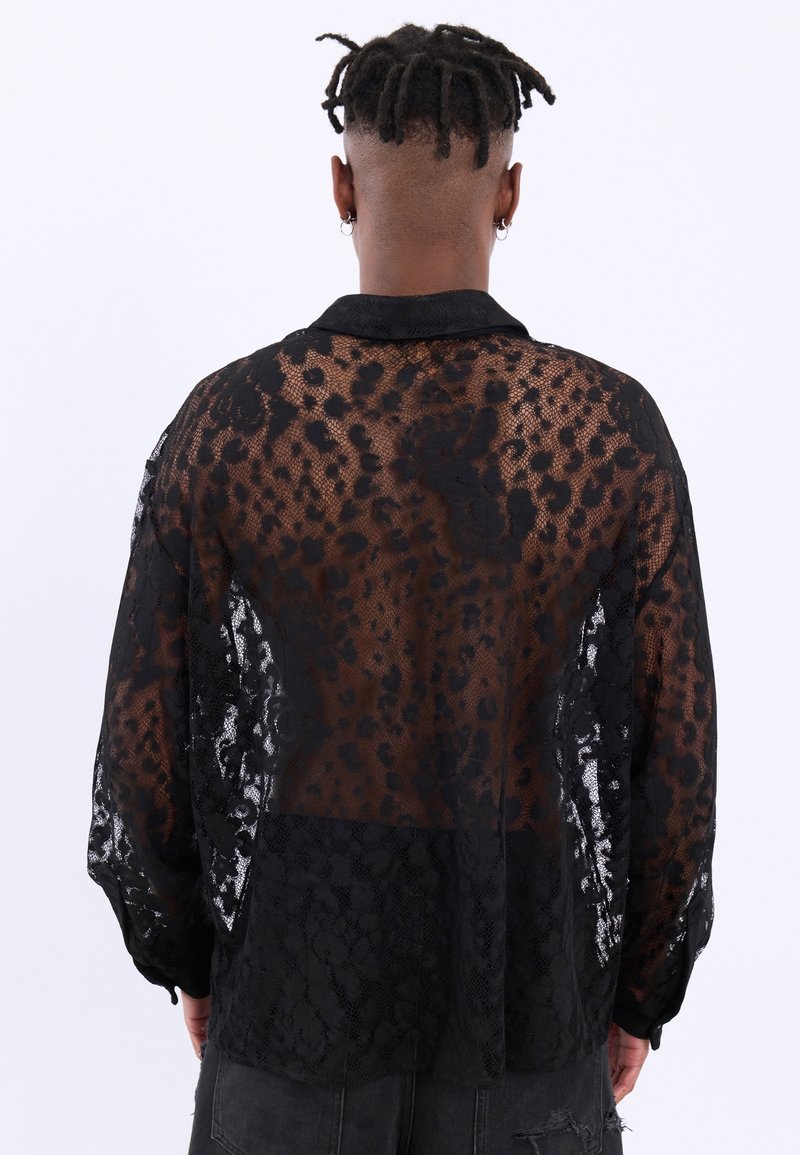 Personne avec des dreadlocks courtes porte une chemise longue en dentelle noire transparente avec un motif floral, vue de dos sur fond blanc.