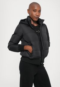 Doudoune noire avec capuche, présentant des sections matelassées, une fermeture éclair à l'avant et des poignets côtelés. Associée à un haut noir et un pantalon.