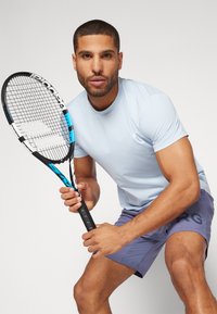 Camisa de manga corta azul claro con cuello redondo; combinada con pantalones cortos grises que tienen un logotipo negro. Sostiene una raqueta de tenis negra y azul.