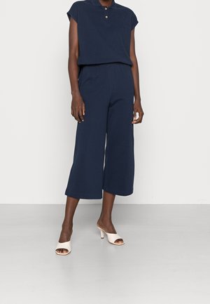 Persoon draagt navy blauwe mouwloze top met kraag en bijpassende brede cropped broek, gecombineerd met witte open-teen sandalen met hak.