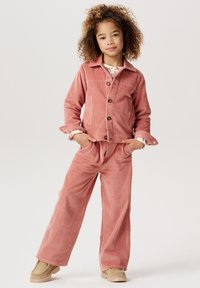 Rosa Cordjacke mit Knöpfen und passender Hose, im lässigen Schnitt und mit gerippter Textur. Das Model trägt ein gemustertes Oberteil darunter.