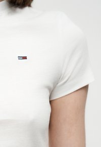 Λευκό στενό πουκάμισο με κοντό μανίκι και μικρό ορθογώνιο λογότυπο σε ναυτικό μπλε, λευκό και κόκκινο στο στήθος, φορεμένο από άτομο με κοντά καστανά μαλλιά.