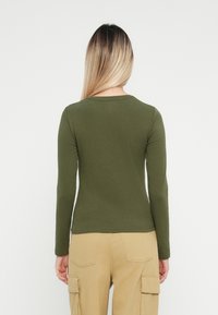 Cămașă lungă cu mâneci verzi-olive, cu un guler rotund, textură fină, design ajustat și un tiv subtil; asociată cu pantaloni cargo de culoare bej deschis.