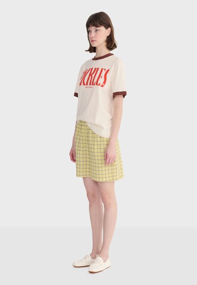 Mladá žena stojaca, oblečená v krémovom tričku s červenou potlačou, žltú károvanú sukňu a biele tenisky, na jednotnom svetlom pozadí.