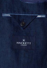 Marineblauer Stoff mit geknöpftem Klappentaschen und aufgenähtem Etikett mit der Aufschrift "Hackett London" in weißem Text unter einem Logo mit gekreuzten Cricket-Schlägern.