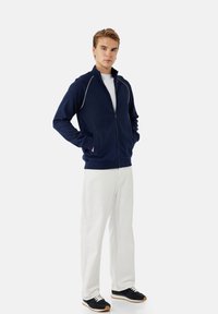 Chaqueta de arriba con cremallera de color marino con ribetes blancos, llevada sobre una camiseta blanca, combinada con pantalones de tonos claros y zapatillas oscuras. Ajuste casual.