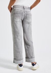 Persona che indossa jeans grigi chiari a gamba larga con orli e sneakers bianche, vista da dietro su uno sfondo bianco uniforme.