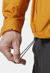 Pomarańczowa lekka kurtka z regulowanym ściągaczem na dolnym brzegu. Prezentuje gładką fakturę oraz dopasowany krój w okolicy nadgarstka. Widziane czarne akcenty przeszycia.