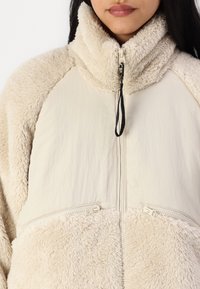 Kremowa kurtka z polaru i gładkiego materiału z wysokim kołnierzem, półzamkiem z przodu oraz dwiema zapinanymi na zamek kieszeniami w talii. Teksturowana i miękka.