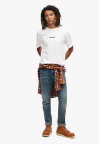 Hvid T-shirt med korte ærmer og "SUPERDRY" logo, blå jeans, orange-brune støvler og en rødternet skjorte bundet om livet.