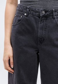 Άτομο που φοράει μαύρο τζιν ψηλόμεσο με ψηλή μέση και σκούρο γκρι μπλουζάκι μαζεμένο μέσα, με το αριστερό χέρι χαλαρά στο πλάι.