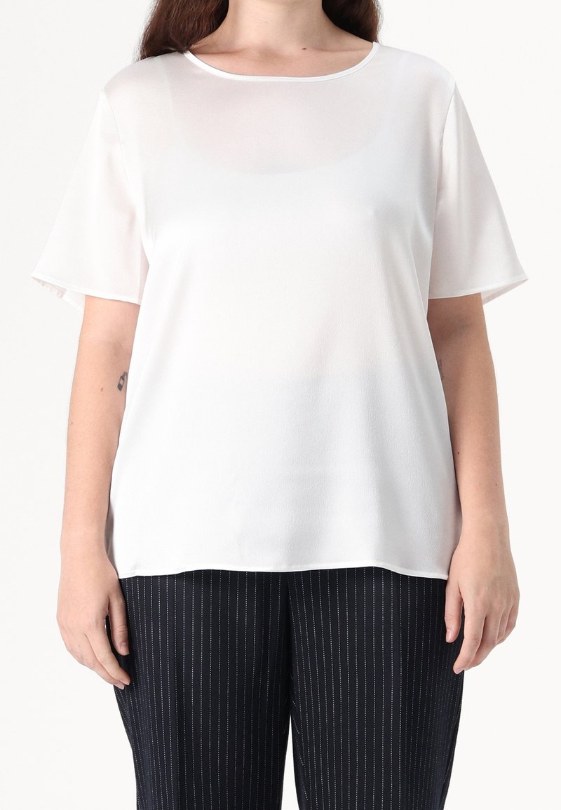Femme portant un chemisier blanc à manches courtes avec un col rond et un pantalon noir à fines rayures, debout devant un fond uni.