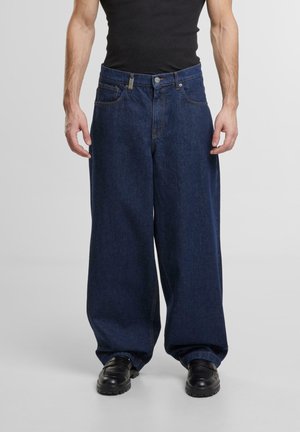 Mann trägt dunkelblaue Jeans mit weitem Bein, ein schwarzes Tanktop und schwarze Schuhe, steht vor einem einfarbigen hellen Hintergrund.