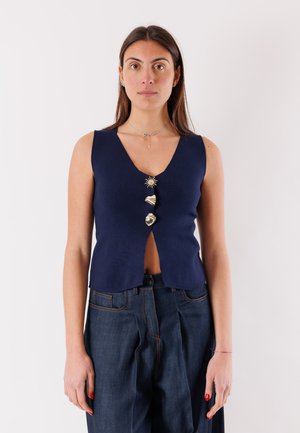 Donna che indossa un top senza maniche blu navy con tre bottoni dorati unici e jeans blu scuro a vita alta, in piedi davanti a uno sfondo semplice.