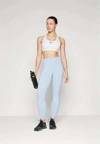 Top desportivo branco com logótipo preto, leggings azul claro e ténis pretos. Segurando uma garrafa de água preta. Design simples, textura suave.
