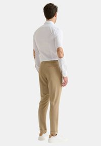 Camicia bianca con toppe color beige sui gomiti, pantaloni beige e sneakers bianche, mostrata da dietro. La camicia ha un colletto classico e un design aderente.