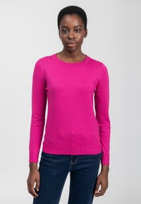 Ung sort kvinde med kort hår iført en lys pink langærmet sweater og mørkeblå jeans, stående mod en neutral baggrund.